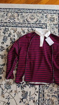 Langärmliges, rot und navy gestreiftes Rugby-Shirt mit weißem Kragen und Etikett, flach auf einem floralen Teppich ausgelegt.