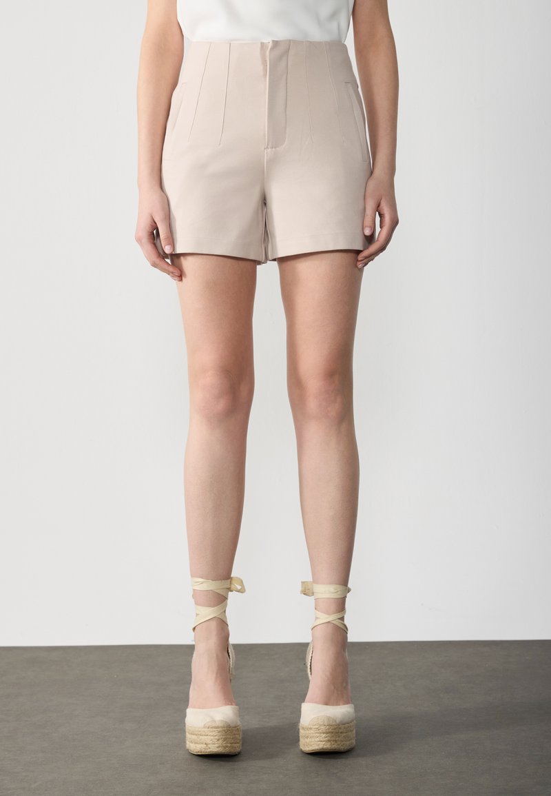 Shorts beiges taille haute avec des plis subtils et des poches latérales, associés à des espadrilles beiges à talons avec des sangles autour des chevilles.