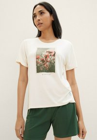 Witte t-shirt met een bloemenprint van roze papavers op een groene achtergrond, gecombineerd met donkergroene shorts. Zacht materiaal, korte mouwen en ronde hals.