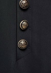 Trois boutons métalliques décoratifs avec des motifs de blason complexes fixés sur un tissu foncé texturé.