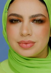 Närbild av en person som bär en ljusgrön strukturerad hijab, med mjuk, neutral sminkning och fylliga läppar, mot en blå bakgrund.