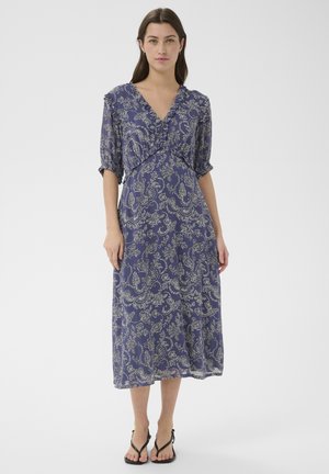 Žena stojí v modrých midi šatách s bielym kvetinovým paisley vzorom, výstrihom do V a rukávmi po lakeť, doplnených o čierne sandále.