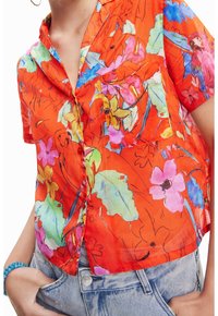 Personne portant une chemise à manches courtes orange vif à motifs floraux roses, verts et bleus, rentrée dans un jean en denim bleu clair.