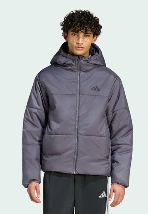 Junger Mann mit lockigem Haar, der eine graue gesteppte Kapuzenjacke und schwarze Adidas-Hosen trägt, steht vor einem schlichten Hintergrund.