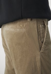 Beige corduroy broek met verticale geribbelde textuur, voorzien van een achterzak en gestikte details. Gedragen met een gedeeltelijk zichtbare donkere top.