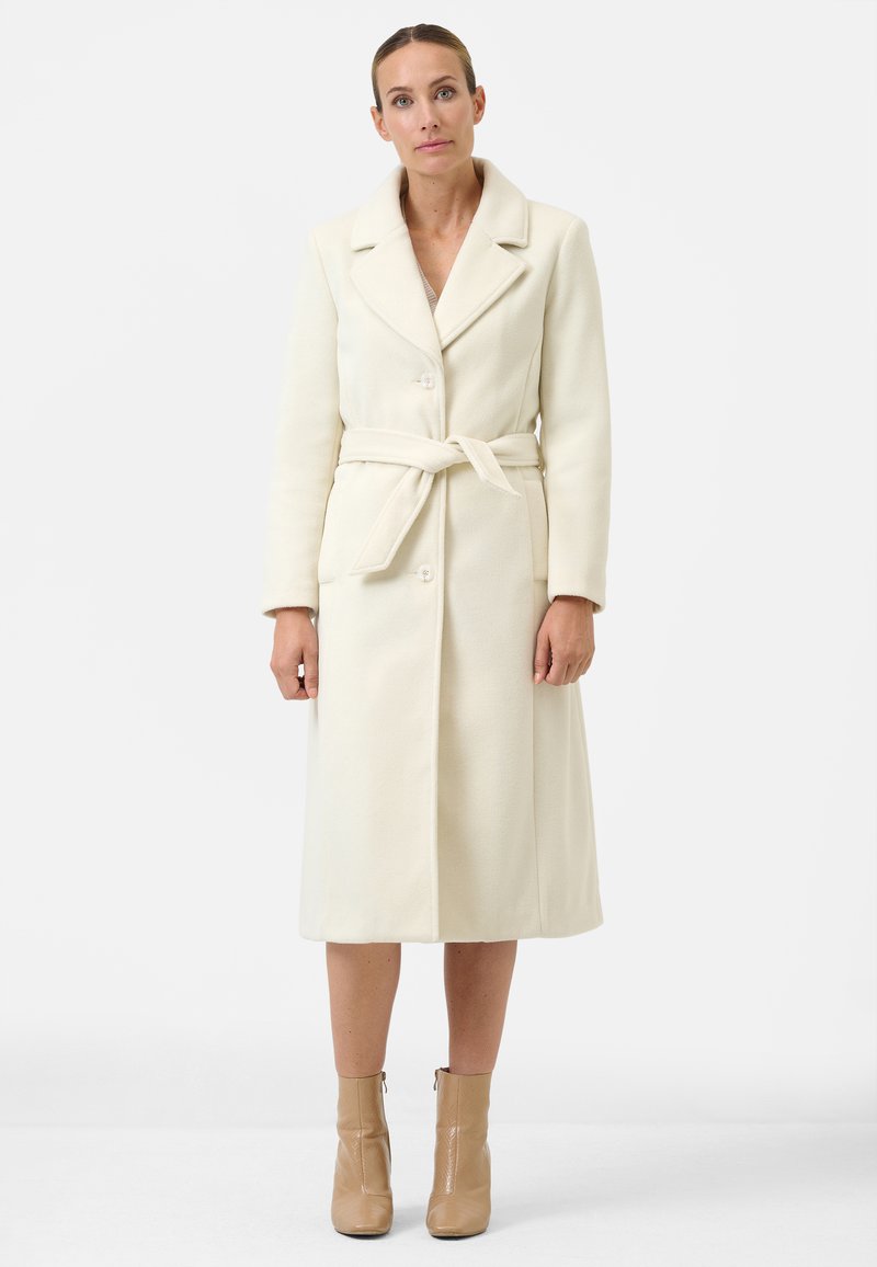 Cream-colored wool coat with a lapel collar, waist tie, and button closure. Features long sleeves and side pockets. Strappy beige boots.
