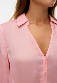 Blouse rose à col en V avec une finition texturée et brillante, dotée d'un col et d'une fermeture à boutons. Tissu doux et léger avec un éclat subtil.