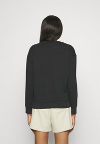 Sweat à col rond en coton noir, avec des poignets et un ourlet côtelés. Présente un petit logo en haut du dos. Associé à un short de couleur claire.