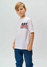 Biała bawełniana koszulka z krótkimi rękawami, z kolorowym graficznym napisem "GOT RIZZ" po lewej stronie. Połączona z niebieskimi dżinsami.