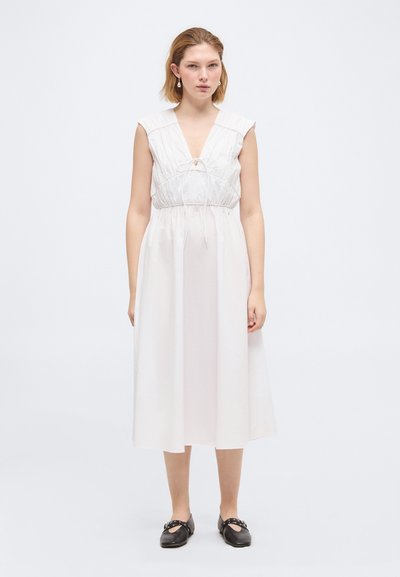 Femme debout portant une robe midi blanche sans manches avec un col en V noué et des chaussures plates noires, sur un fond blanc uni.
