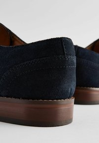 Zapatos de ante azul marino con talones redondeados, que presentan costuras visibles y suelas de madera marrón para contraste. Una textura sutil realza el diseño.