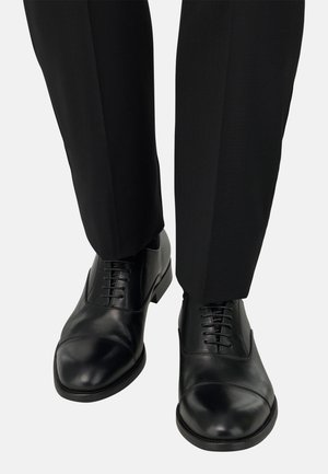 Zapatos de vestir de cuero negro con punta redondeada y diseño clásico de cordones, combinados con pantalones entallados negros. Textura suave, sin patrones visibles.