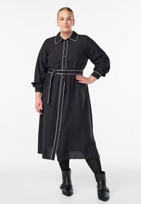 Žena stoji samouvjereno, odjevena u crnu haljinu dugih rukava sa pojasom i bijelim obrubom, crne tajice i crne čizmice do gležnja, nasuprot bijeloj pozadini.