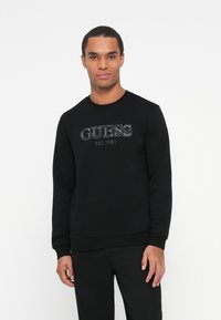 Fiatal férfi fekete pulóverben, amelyen a "GUESS EST. 1981" logó látható, fehér háttér előtt áll, előre nézve, semleges arckifejezéssel.