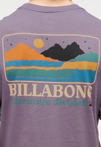 Τοπίο με βουνό και λίμνη με αστέρια και ήλιο γραφικά σε μωβ μπλουζάκι, με το κείμενο «BILLABONG Adventure division» από κάτω.