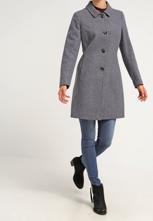 Frau trägt einen kniehohen grauen Knopfmantel, blaue enge Jeans und schwarze Stiefeletten, geht mit einem Bein vor das andere gekreuzt.