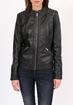 Mujer con una chaqueta de cuero negra ajustada con cremallera frontal y bolsillos laterales con cremallera, combinada con vaqueros azul oscuro sobre un fondo blanco.