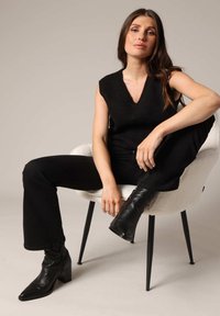 Débardeur noir à côtes sans manches avec un décolleté en V, associé à un jean noir évasé et des bottes à talons pointus noires, assis sur une chaise d'accent blanche.