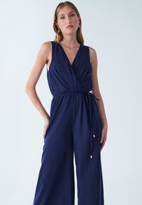 Temno modra pajkica brez rokavov ima globok V-izrez, obliko ovitka, pasni trakec in široke hlačnice, izdelana iz mehke, teksturirane tkanine.