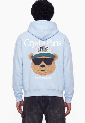 Person wearing light blue hoodie with a bear face wearing sunglasses and a cap, text "Crystal Paris" and "Summer 2026 © CP" on the back.
