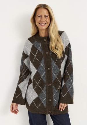 Smilende kvinde med langt blondt hår iført en brun og grå argyleknappet cardigan og mørke bukser mod en ensfarvet baggrund.