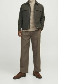 Completo da uomo composto da una giacca grigia scura a quadri con due tasche frontali, un dolcevita beige, pantaloni marroni plissettati e scarpe marroni.