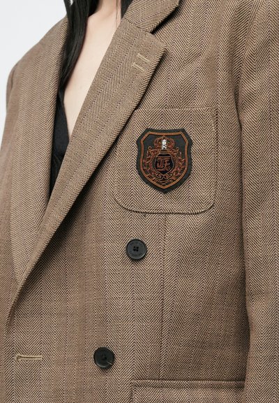 Sacou din lână în carouri maro, având un patch brodat negru pe buzunarul de la piept, două butoane negre și reveruri structurate.