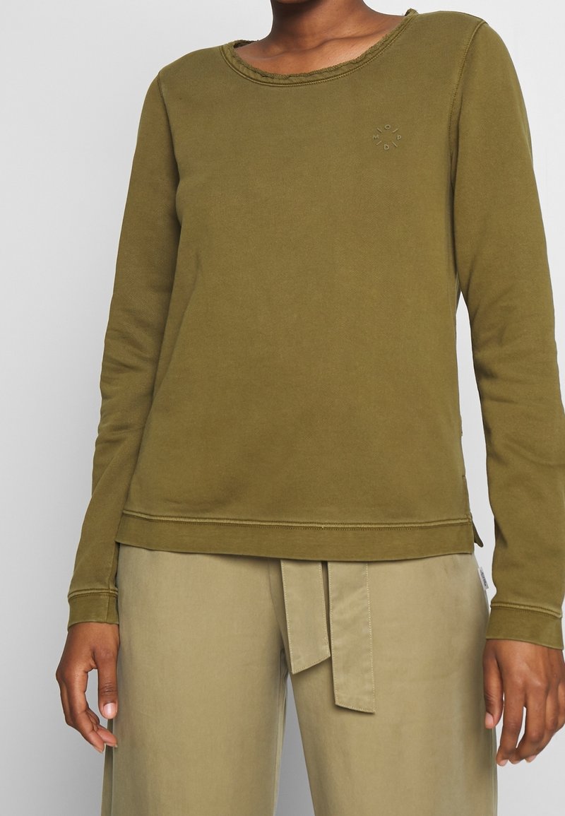 Persoon in een olijfgroene lange mouwen shirt en beige broek met een gestrikte riem, staande met ontspannen handen langs hun zij.