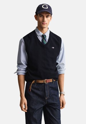 Mies käyttää laivastonsinistä lippalakki, jossa on "G", vaaleansinistä nappikauluspaitaa, laivastonsinistä neuleliiviä, raidallista solmiota, ruskeaa vyötä, tummaa farkkua ja hopeakelloa, seisoo valkoista taustaa vasten.