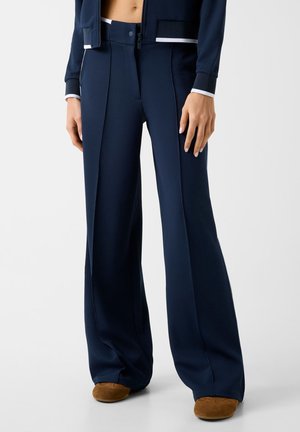 Marineblaue Hosen mit weitem Bein und strukturierter Stoffqualität, kombiniert mit braunen Schuhen und einer passenden marineblauen Jacke, die eine freiliegende Taille zeigt.