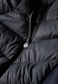 Chaqueta acolchada azul marino con un diseño elegante, que presenta un cierre de cremallera y una lengüeta metálica. El material parece ligero y tiene una textura acolchada.