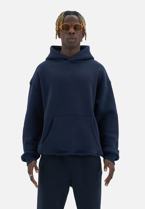 Navy blue oversized hoodie made of soft fabric, featuring a kangaroo pocket and front logo. Model wears matching navy pants and orange glasses.