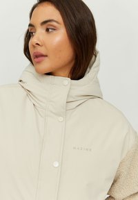 Beige padded jacket with a high collar, featuring a soft textured sleeve, snap buttons, and embroidered brand name "MAZINE" on the front.