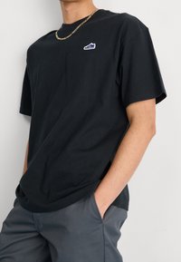 Camiseta de algodón negra con un pequeño logo de zapatilla azul y blanco en el pecho, combinada con pantalones grises de corte ajustado.