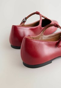 Zapatos de cuero rojo con punta redondeada y tira en el tobillo, vistos desde la parte posterior, descansando sobre un fondo neutral.