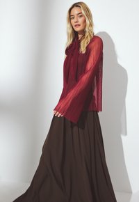 Prosojna burgundska bluza s pregibano teksturo in detajlom na vratu v obliki pentlje, v kombinaciji z dolgo rjavo krilo, ki ima tekoč dizajn in gladko tkanino.