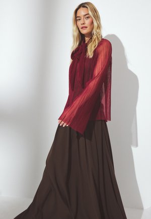 Blusa burdeos transparente con textura plisada y detalle de lazada en el cuello, combinada con una larga falda marrón de diseño fluido y tejido suave.