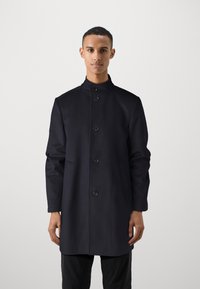 Cappotto blu navy con design strutturato, realizzato in tessuto testurizzato con colletto alto, sei bottoni e tasche laterali. Aspetto semplice e moderno.