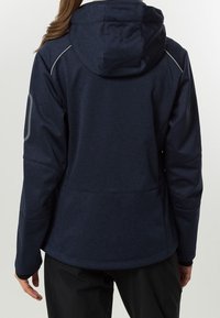 Chaqueta con capucha de color azul marino que presenta una textura suave, costuras en ángulo, acentos reflectantes y un dobladillo ligeramente curvado en la parte trasera.