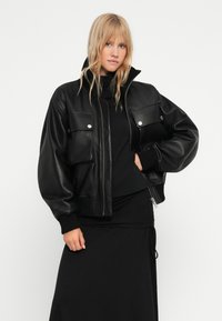 Crna kožna bomber jakna s oversized fitom, rebrastim manžetama, dva džepa na prsima i visokim okovratnikom. Zatvaranje na zip i glatka završna obrada.