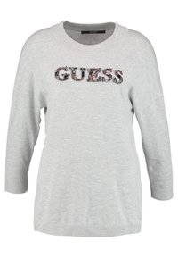 Světle šedý svetr z měkké látky, s kulatým výstřihem a 3/4 rukávy. Má texturované logo "GUESS" zdobené barevnými akcenty.