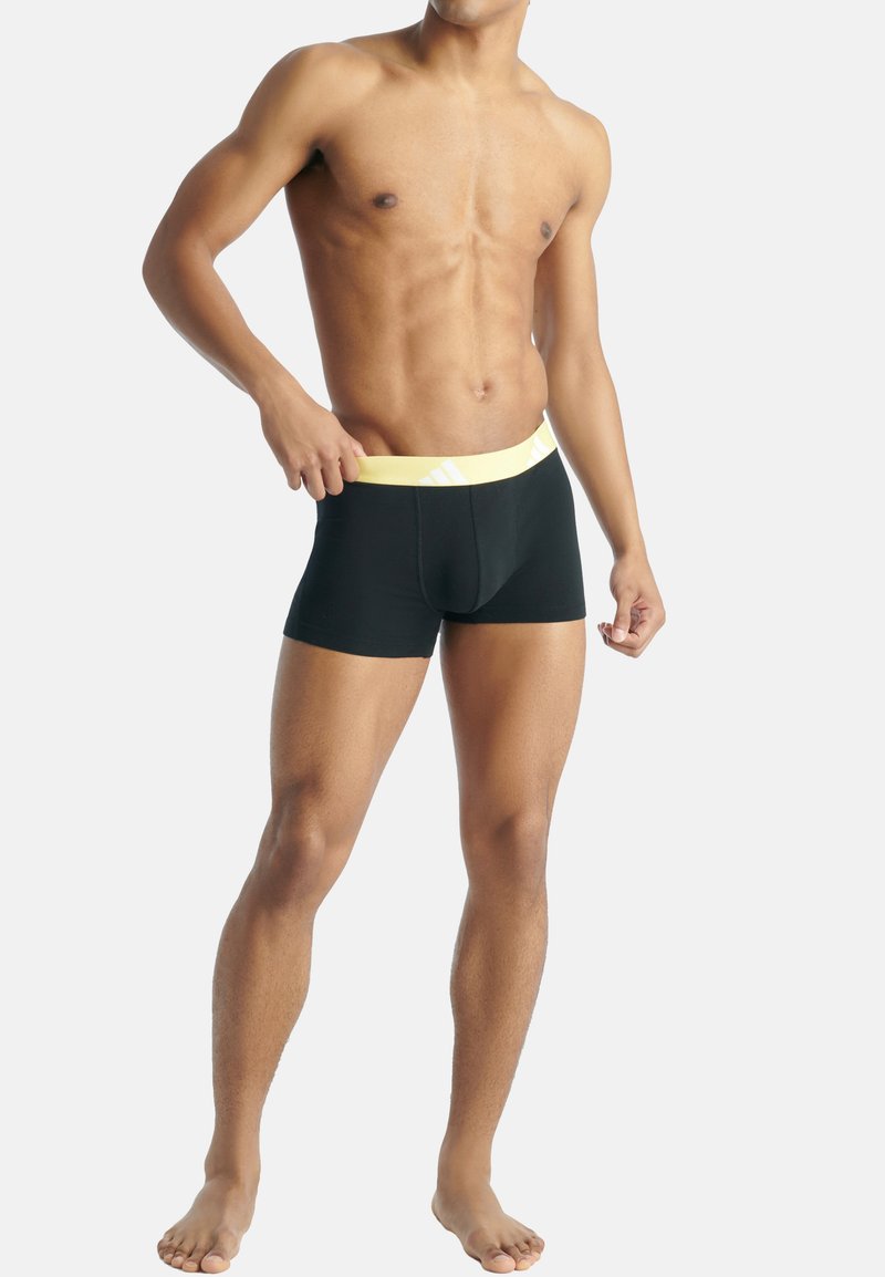Schwarze Boxershorts mit gelbem elastischem Bund, der ein gestreiftes Design aufweist. Glattes Material, figurbetonte Form und Mittellänge bis zum Oberschenkel.