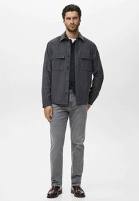 Man in donkergrijze button-up jasje over een zwart shirt, lichte grijze jeans en donkerbruine loafers, staande voor een effen witte achtergrond.