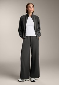 Geacă zip-up de culoare gri închis, cu un model striat și accente elastice, combinată cu pantaloni largi asortați și un top alb ajustat. Tenisul alb completează ținuta.