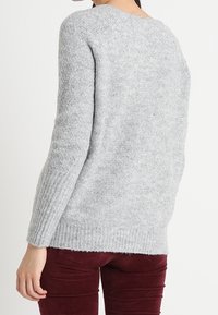 Pull en maille gris avec une coupe décontractée, des poignets et un ourlet côtelés, et une texture douce. Porté avec un pantalon bordeaux, affichant un design simple et décontracté.
