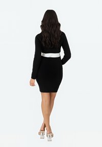 Czarna, prążkowana sukienka bodycon z długimi rękawami i akcentem w postaci białego pasa. Sukienka jest dopasowana, sięgająca powyżej połowy uda. Ubrano przezroczyste obcasy.