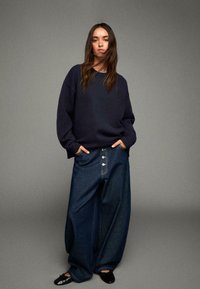 Merik Bleu oversize neule, yhdistettynä leveälahkeisiin tummiin farkkuihin, joissa on nappisuljenta. Malli seisoo kädet taskuissa, mustat loafersit näkyvissä.