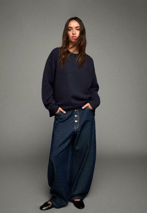 Pull oversized bleu marine associé à un pantalon en denim noir à jambes larges avec un devant à boutons. Le mannequin se tient avec les mains dans les poches, des mocassins noirs visibles.