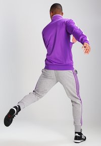 Lila träningsjacka med vita accenter, grå joggingbyxor med lila ränder, och svarta träningsskor. Modellen står i en dynamisk pose.