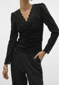 Schwarze Spitzenbluse mit gewelltem Halsausschnitt und gerafftem Vorderteil; verfügt über lange Ärmel mit Rüschensäumen, kombiniert mit gestreiften schwarzen Hosen.